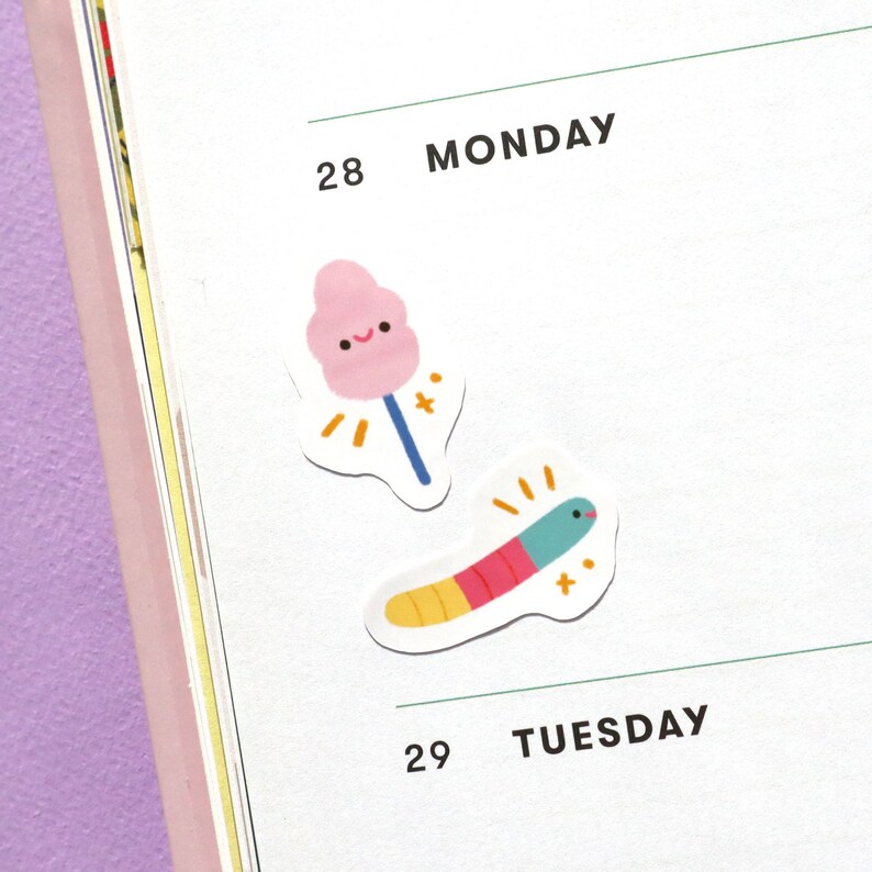May include: Two stickers on a white planner page. The top sticker is pink and shaped like a cotton candy cloud with a blue stick. The bottom sticker is a colourful worm with a green head and yellow, red, and blue stripes.