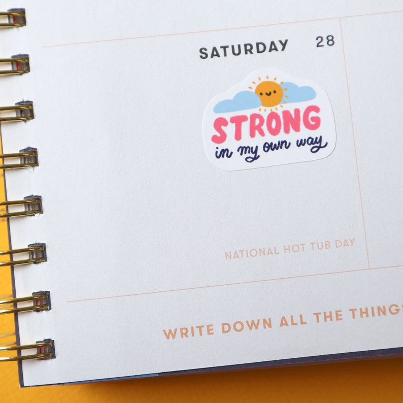 May include: A white sticker with a yellow sun, blue clouds, and pink text that reads "STRONG in my own way". The sticker is on a white page with the text "SATURDAY 28" and "NATIONAL HOT TUB DAY" printed below.