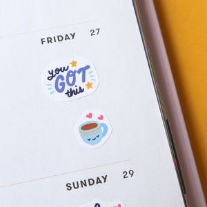 May include: Two motivational stickers for a planner or journal. One sticker is white with blue and purple lettering that says "you got this" with two stars. The other sticker is white with a blue and white cartoon mug with a brown liquid inside and two pink hearts above it.