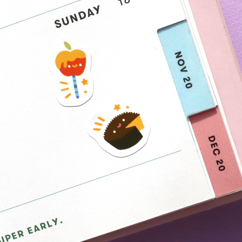 May include: Two colourful stickers featuring a cartoon apple on a stick and a cartoon cupcake with a smiling face. The stickers are on a white planner page with the word "SUNDAY" and the dates "NOV 20" and "DEC 20" visible.