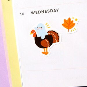 May include: A sticker featuring a cartoon turkey with a ghost riding on its back. The turkey is brown with orange feathers. The ghost is white with a smiling face. There is also a sticker of an orange leaf with a star.