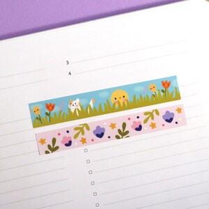 May include: Two decorative washi tape strips. One features a cartoon scene with a cat, sun, and flowers against a blue sky. The other has a pink background with flowers and leaves. Both are on a lined paper.