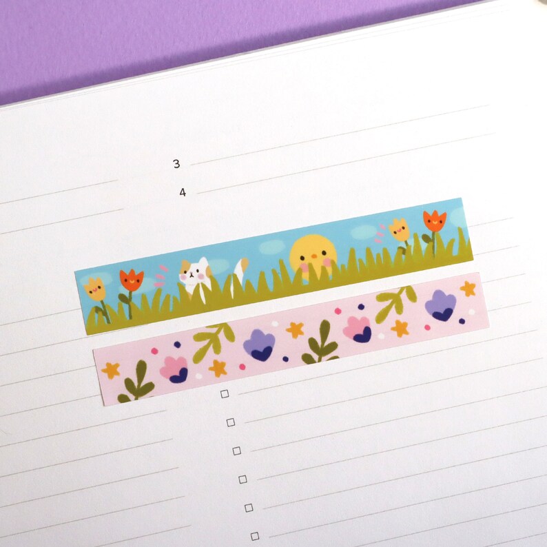 May include: Two decorative washi tape strips. One features a cartoon scene with a cat, sun, and flowers against a blue sky. The other has a pink background with flowers and leaves. Both are on a lined paper.
