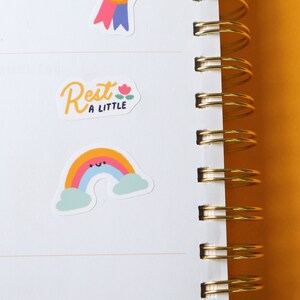 May include: A white notebook with a gold spiral binding. The notebook has a sticker of a rainbow with a smiley face and a sticker that says "Rest a little" with a small flower.