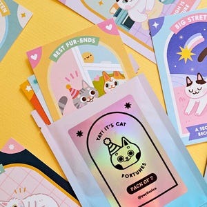 May include: A collection of colorful cat-themed fortune cards and a packet of seven fortunes. The cards feature cartoon cats with various designs and phrases like "Best Fur-ends" and "Big Stretch." The packet has a holographic label with a cat illustration and the text "Yay! It's Cat Fortunes."