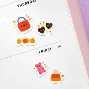 May include: A set of five Halloween-themed stickers featuring a jack-o'-lantern bucket, two chocolate hearts, a candy bar, a pink gummy bear, and a candy corn. The stickers are on a white background.