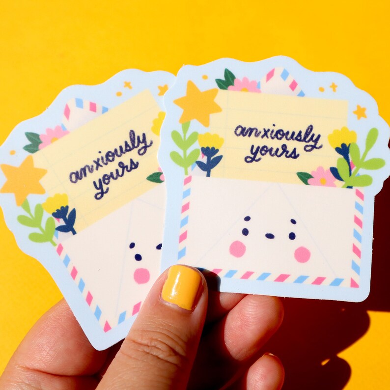 Pode incluir: Dois adesivos de envelope azul e branco com um rosto de desenho animado fofo e o texto "anxiously yours" em escrita cursiva preta. Os adesivos s&atilde;o decorados com estrelas amarelas, folhas verdes e flores rosa e amarelas.