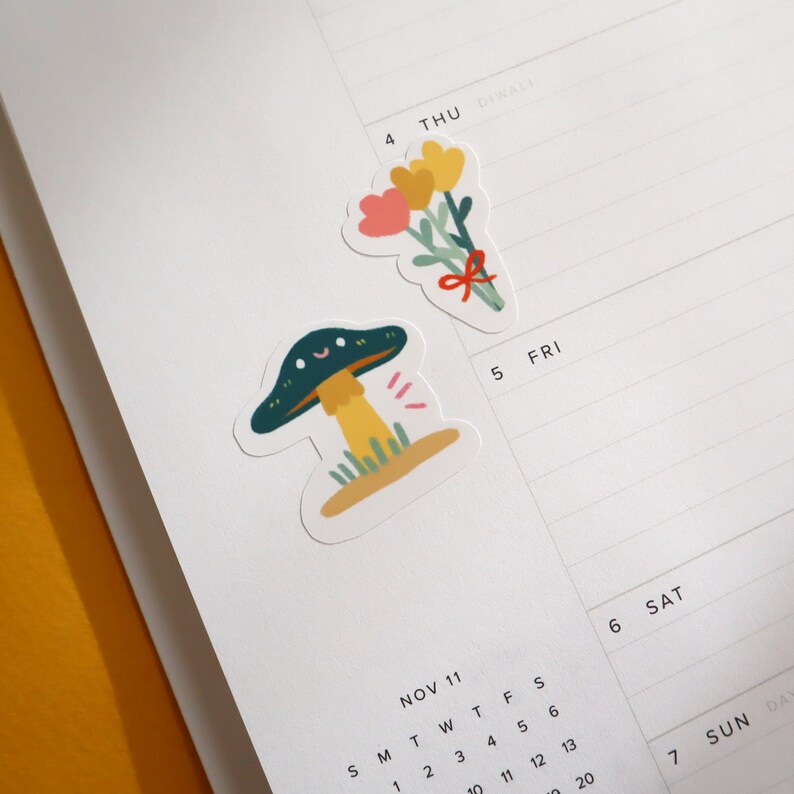 May include: Two colorful stickers on a white calendar page. One sticker features a green mushroom with a yellow stem and a smiling face. The other sticker features a bouquet of yellow and pink flowers with a red ribbon.