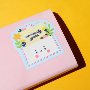 Pode incluir: Um adesivo com um envelope de desenho animado com um rosto bonito e uma nota dentro que diz "anxiously yours". O envelope &eacute; decorado com flores e uma estrela.