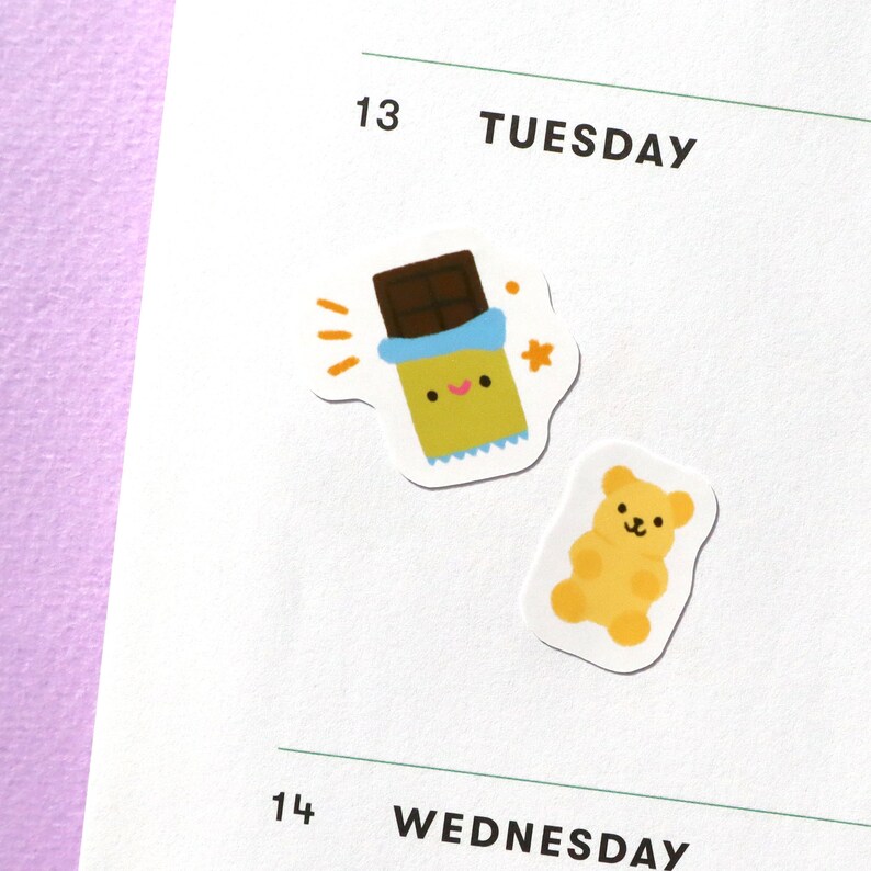May include: Two stickers, one with a cartoon chocolate bar with a smiling face and the other with a cartoon yellow bear. The stickers are on a white page with the text "13 TUESDAY" and "14 WEDNESDAY" above and below the stickers.