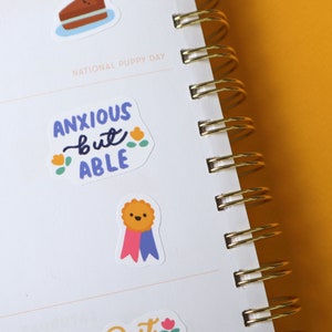 May include: A white planner with a gold spiral binding. The planner has stickers on it that say "National Puppy Day" and "Anxious but Able". One sticker features a cartoon dog wearing a blue and pink ribbon.