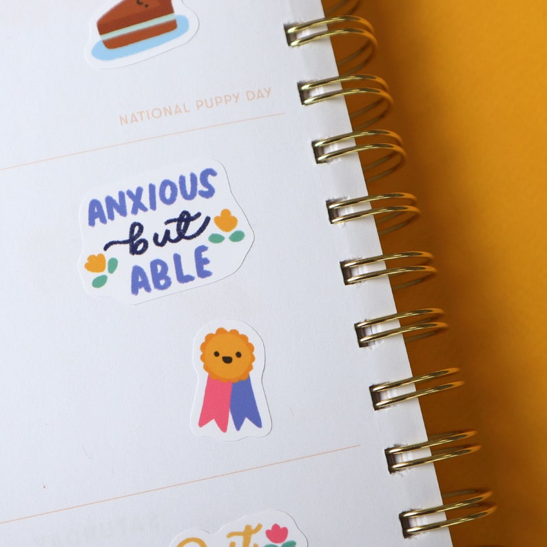 May include: A white planner with a gold spiral binding. The planner has stickers on it that say "National Puppy Day" and "Anxious but Able". One sticker features a cartoon dog wearing a blue and pink ribbon.
