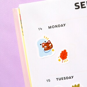 May include: A white ghost holding a brown cat with a yellow star sticker on a white planner page with the text "MONDAY" and "TUESDAY" on the page.