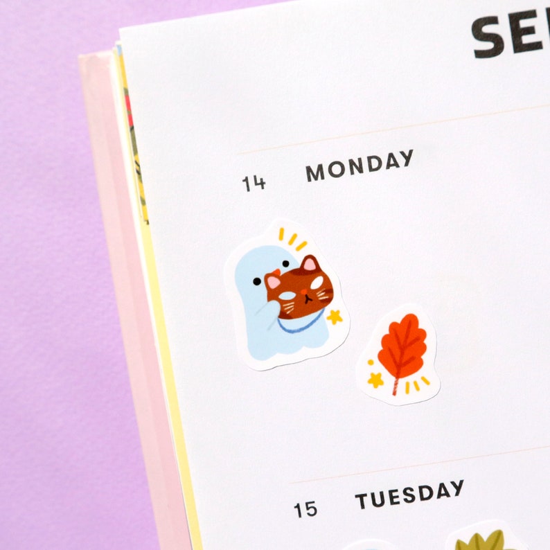 May include: A white ghost holding a brown cat with a yellow star sticker on a white planner page with the text "MONDAY" and "TUESDAY" on the page.