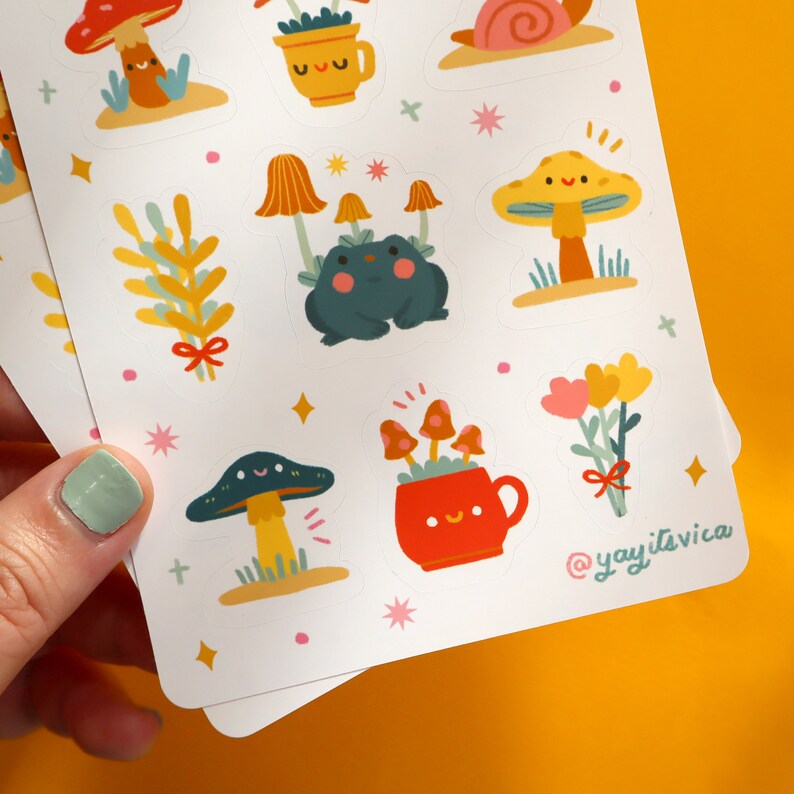 May include: A sheet of colorful stickers featuring various cute illustrations of mushrooms, a frog, a snail, flowers, and plants. The stickers are on a white background with a light blue border. The text "@yayitsvica" is printed in the bottom right corner.