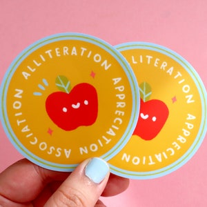 May include: Two yellow and blue round stickers with a red apple and the text "Alliteration Appreciation Association" on them.