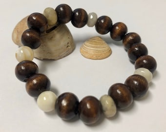 Puede incluir: Pulsera de cuentas con cuentas de madera marrón oscuro y cuentas redondas blanquecinas alternadas. La pulsera se exhibe sobre una superficie blanca con conchas marinas en el fondo. Las cuentas tienen aproximadamente 1,3 cm de diámetro.