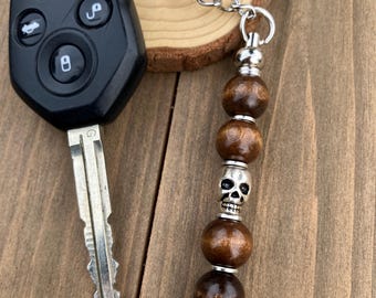 May include: A black car key with a silver key ring attached to a beaded keychain. The keychain features dark brown wooden beads, a silver skull bead, and silver accents. The key fob has red and white buttons.
