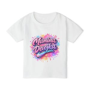May include: White t-shirt featuring the text "Mamina Devojka" in a colourful, stylised font. The text is set against a splash of pink, blue, and yellow paint, creating a vibrant design.