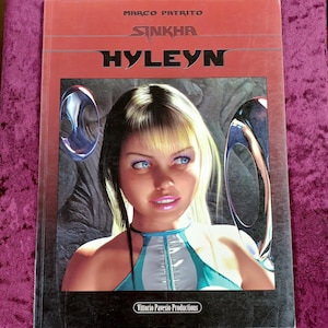 以下が含まれることがあります： マルコ・パトリト著のグラフィックノベル「Sinkha Hyleyn」。金髪で青い目の人物がターコイズブルーと白のトップスを着用している肖像画が描かれている。表紙には金属的な円形の形状と「Vittorio Pavesio Productions」という文字が含まれている。
