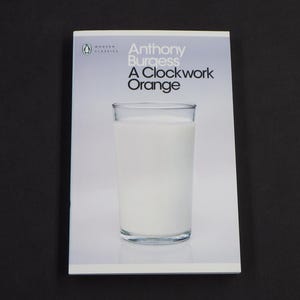 May include: A paperback book cover with a white background and black text. The title is "A Clockwork Orange" by Anthony Burgess. The cover features a glass of milk.