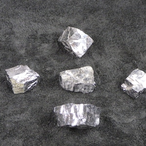 May include: Five silver-colored, irregularly shaped metal chunks on a dark gray surface.