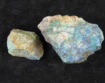 May include: Two rough-cut mineral specimens with a mix of colors. One is a smaller, rectangular piece with green and brown hues. The other is larger, with blue, green, and white colors. Both are on a black background.