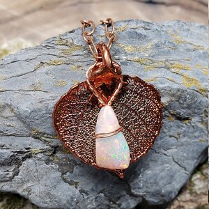 May include: A copper pendant necklace featuring a leaf-shaped design with intricate veining. The pendant holds a white opal gemstone, wrapped in copper wire. The necklace is displayed on a gray stone surface.