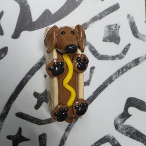 May include: A whimsical, handmade polymer clay figurine of a dachshund hot dog. The brown dog has black paw prints and a black nose, nestled in a bun with yellow mustard. A fun, unique piece.