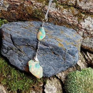 May include: A silver chain necklace with two iridescent opal stones hanging from it. The necklace is displayed on a piece of tree bark with moss.