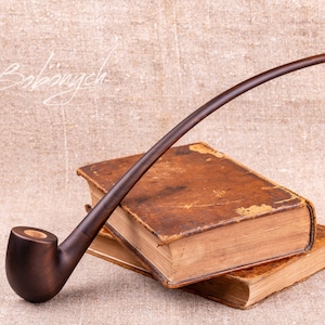 May include: A long, curved wooden pipe resting on top of two antique books with worn leather covers. The pipe is dark brown and has a simple bowl.