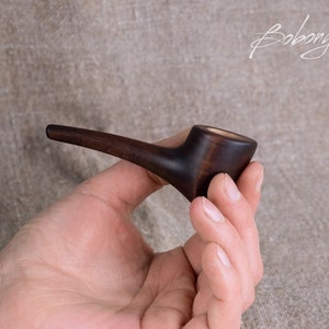 May include: A dark brown wooden pipe with a rounded bowl and a long, curved stem. The pipe is held in a person's hand.
