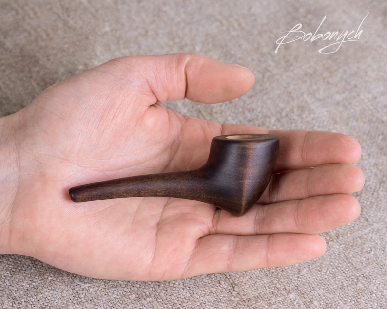 May include: A dark brown wooden pipe with a rounded bowl and a long, curved stem. The pipe is resting in the palm of a hand.