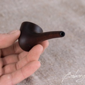 May include: A small, dark brown wooden pipe with a rounded bowl and a long, thin stem. The pipe is held in a hand.