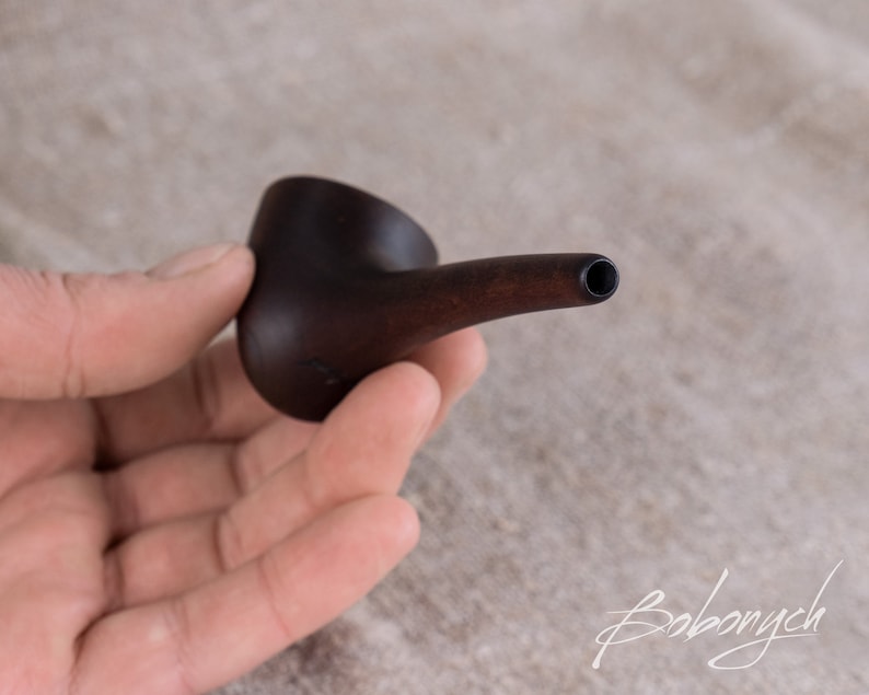 May include: A small, dark brown wooden pipe with a rounded bowl and a long, thin stem. The pipe is held in a hand.