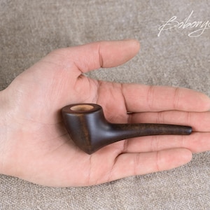 May include: A small, dark brown wooden pipe with a rounded bowl and a long, thin stem. The pipe is held in a person's hand.