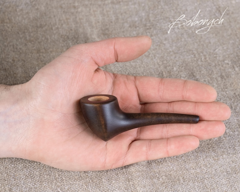 May include: A small, dark brown wooden pipe with a rounded bowl and a long, thin stem. The pipe is held in a person's hand.