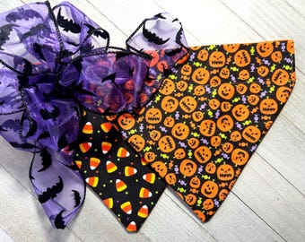 May include: A pet bandana featuring a black fabric with an orange jack-o'-lantern and candy pattern, paired with a second black fabric section displaying a candy corn print. A large, sheer purple bow with a black bat pattern is attached to the top.