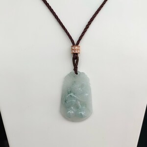 Op de afbeelding: Een jade hangerketting met een gegraveerd ontwerp van een draak. De hanger is lichtgroen en hangt aan een bruin koord met een klein, met diamanten bezet sluiting.