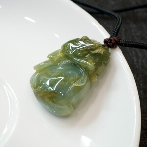 Può includere: Un ciondolo in giada verde intagliato a forma di creatura mitologica, appeso a un cordoncino nero. Il ciondolo è su un piatto bianco.