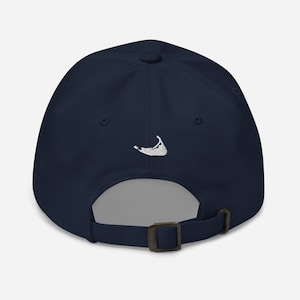 Può includere: Un berretto da baseball blu navy con un contorno di un'isola ricamato in bianco. Il berretto ha un cinturino regolabile con una fibbia in metallo.
