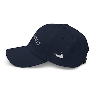 Può includere: Cappellino da baseball blu navy con la scritta "Nantucket" ricamata in bianco sul davanti. Un contorno bianco dell'isola è ricamato sul lato. Il cappellino ha una visiera curva e un cinturino regolabile.