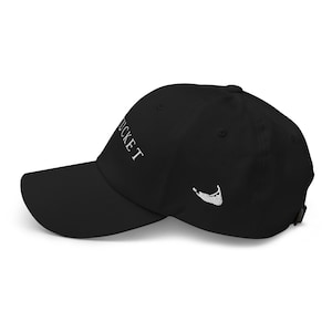 May include: Black baseball cap with the word "Nantucket" embroidered in white on the front. A white outline of the island is embroidered on the side. The cap has a curved brim and an adjustable strap.