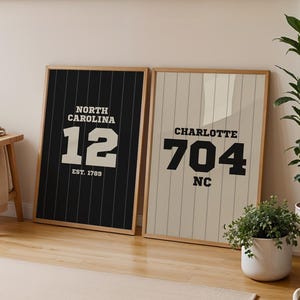 Puede incluir: Dos impresiones enmarcadas se apoyan contra una pared. Una es negra con texto blanco que dice "NORTH CAROLINA 12 EST. 1789". La otra es beige con texto negro que dice "CHARLOTTE 704 NC". La habitación tiene un taburete de madera y plantas.