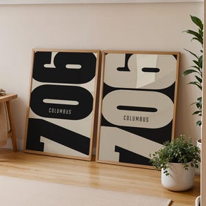 May include: Two framed art prints with the text "907 COLUMBUS" in a bold, modern font. The prints are in a room with a wooden stool, a rug, and potted plants. The color scheme is neutral with black and beige accents.