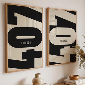 May include: Two framed art prints with a minimalist design. Each print features the word "Orlando" in a black and cream color scheme. The prints are displayed on a white wall above a wooden table with decorative items.