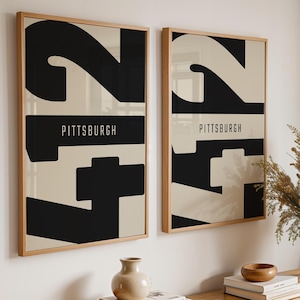 May include: Two framed art prints with abstract black and cream designs and the word "PITTSBURGH" in white. The prints are displayed on a white wall above a wooden table with decorative items, including a vase of dried flowers.