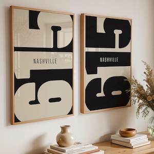 May include: Two framed art prints with a minimalist design. Each print features the word "Nashville" in white text on a black background, with large, abstract shapes in black and cream. The frames are light wood.