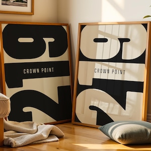 Op de afbeelding: Twee ingelijste kunstprints met een minimalistisch ontwerp. Elke print heeft een groot nummer "2" in zwart en wit, met "CROWN POINT" in witte tekst. De frames zijn gemaakt van lichtbruin hout. Een kleine ingelijste foto van palmbomen hangt boven een van de prints.