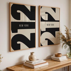 Puede incluir: Dos láminas enmarcadas con un diseño minimalista. Cada lámina presenta un gran número negro "2" sobre un fondo crema, con un rectángulo negro en el medio que dice "NEW YORK" en blanco. Los marcos son de color marrón claro.